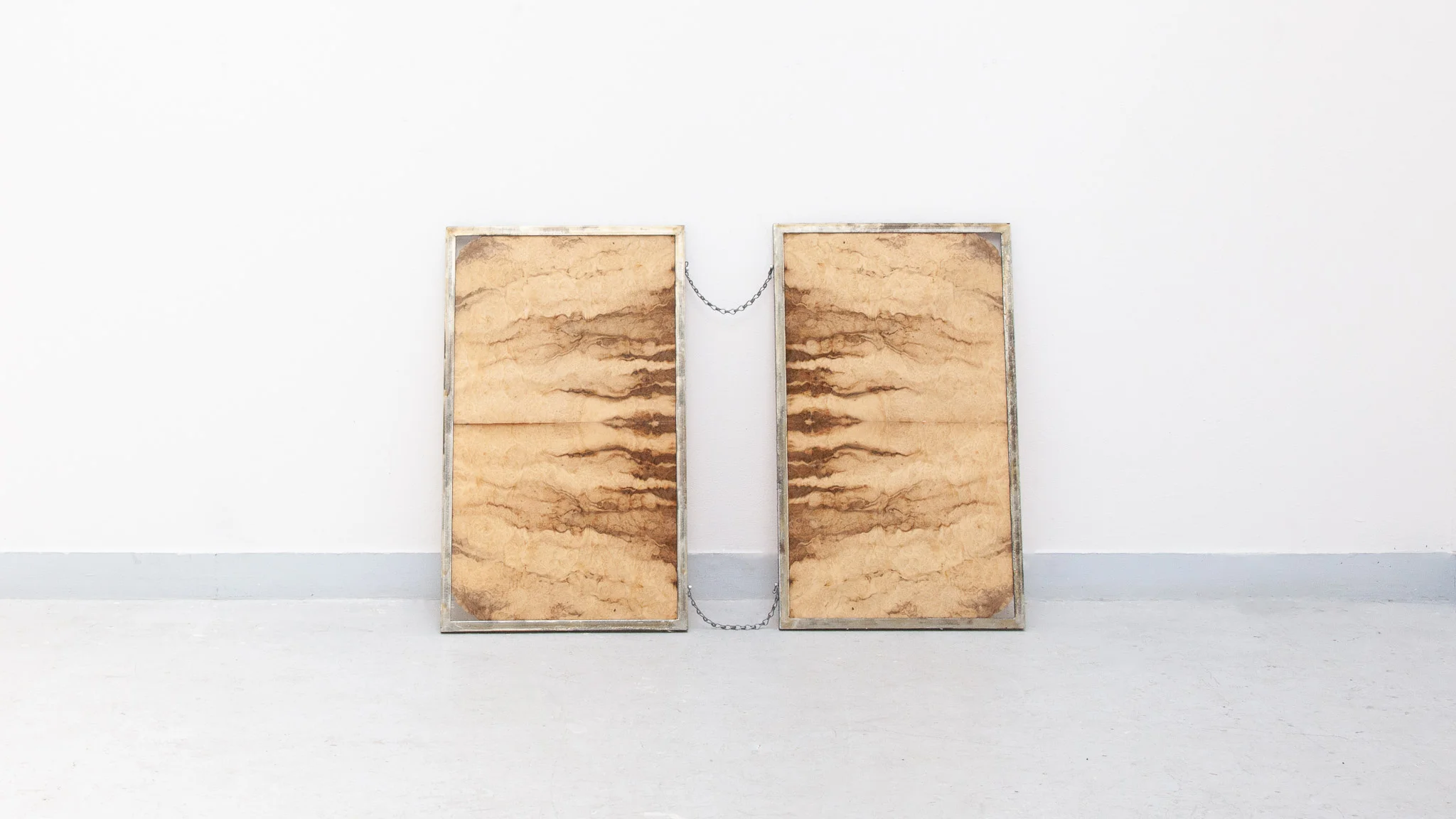 Minimal installation with two rectangular panels featuring marbled, organic patterns, placed side by side on the floor and connected by a small chain against a white gallery wall.