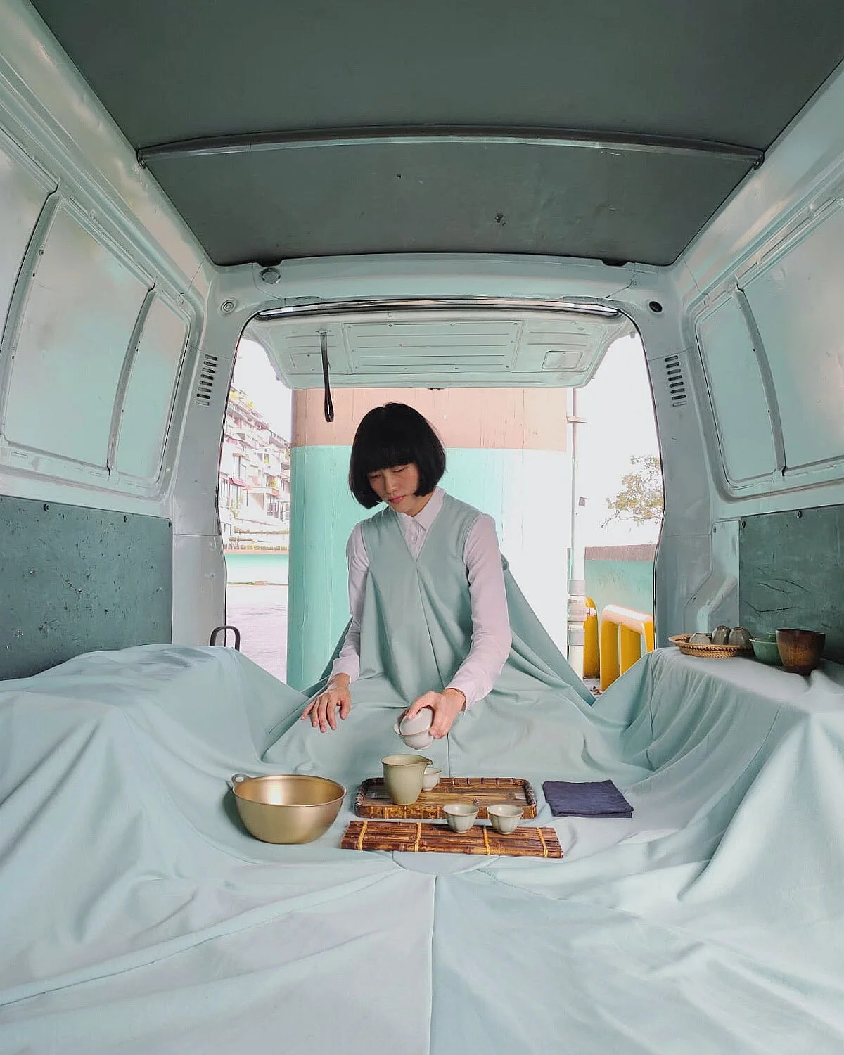 Person performing a tea ceremony inside the back of a van, surrounded by soft draped fabric, carefully pouring tea into small cups arranged on a tray in a calm, intimate setting.