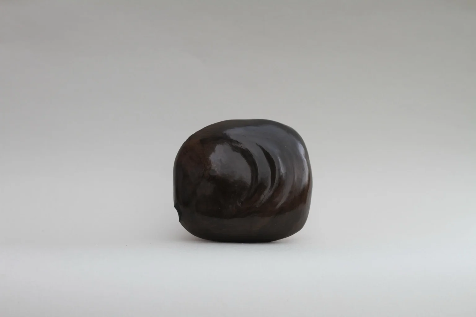 Dark organic sculptural form with a smooth, rounded surface and subtle indentations, resembling a natural or bodily shape, set against a neutral background.