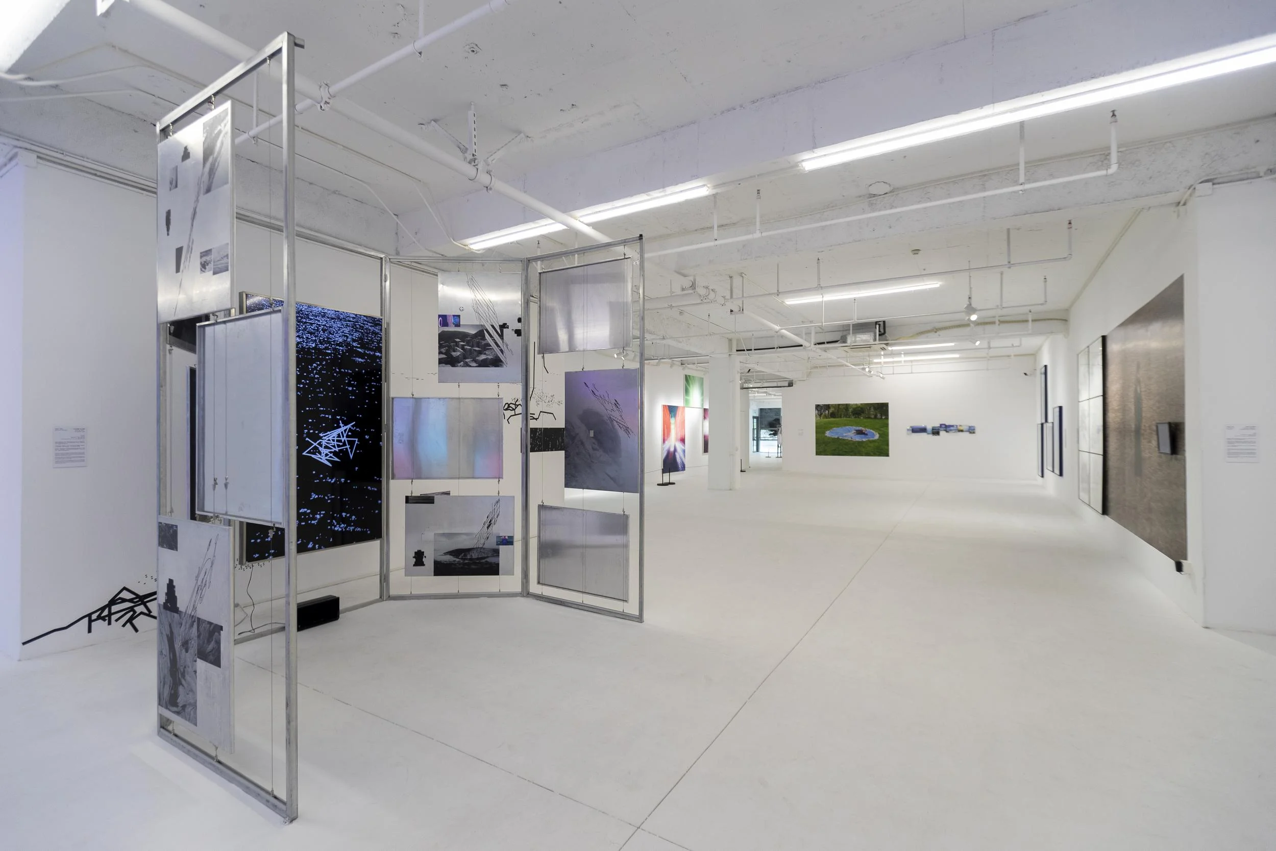 Contemporary gallery installation in a bright white exhibition space, featuring modular metal structures with layered images, digital screens, and large-scale artworks displayed across the room.