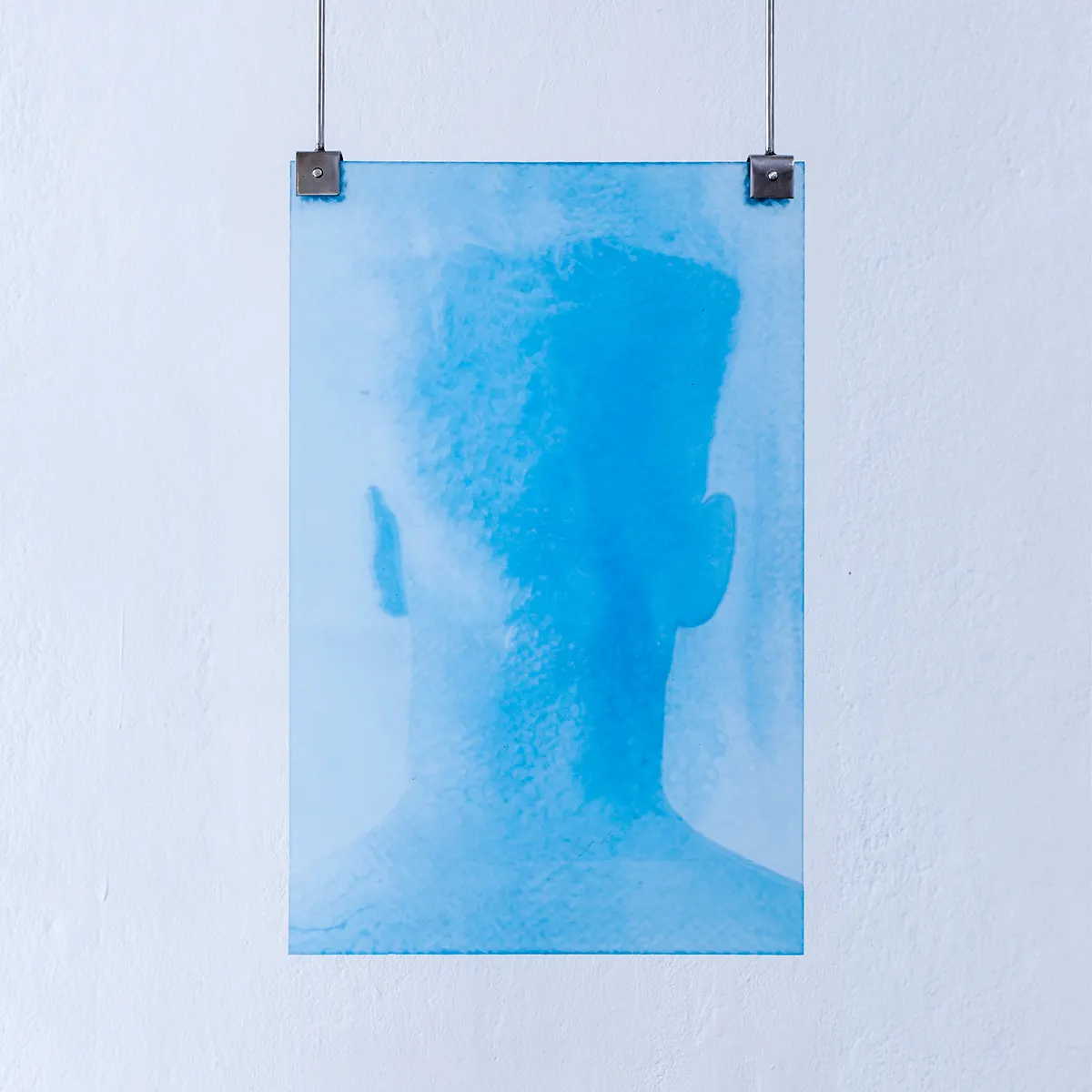 Translucent blue artwork depicting a blurred human head silhouette, suspended from metal clips against a white wall, creating a soft, atmospheric portrait-like image.