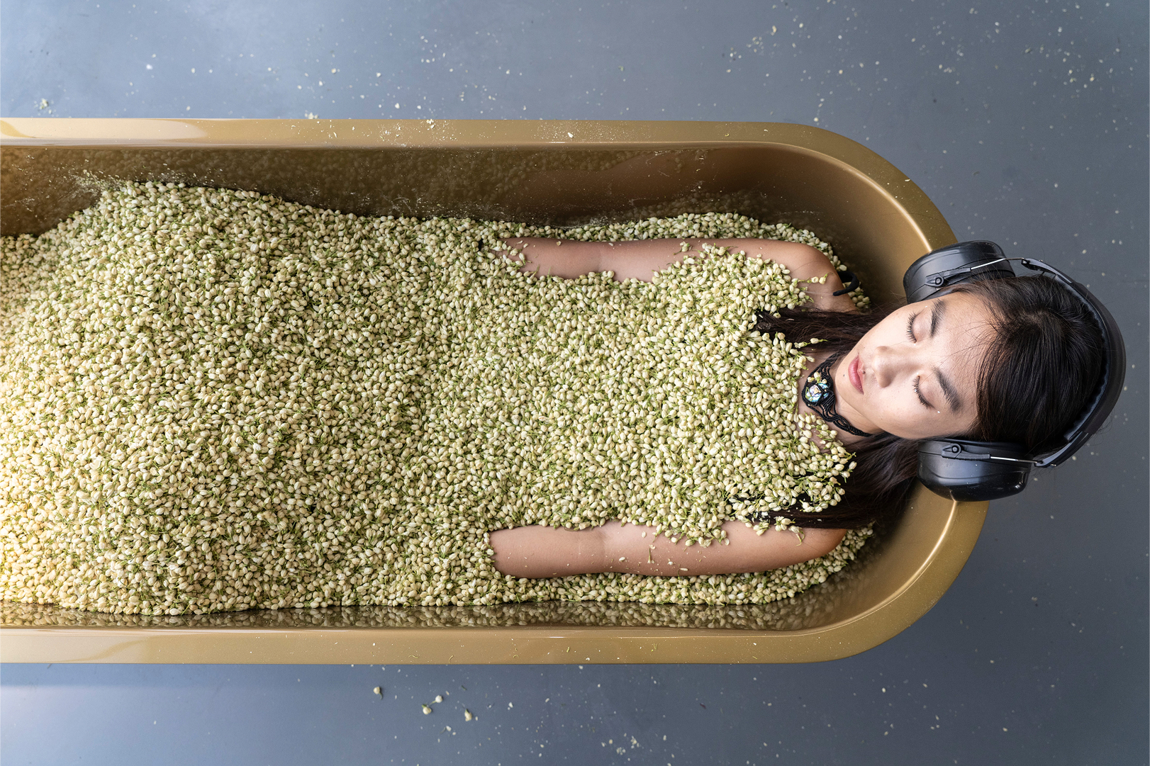Performer submerged in hemp seeds in gold bathtub wearing headphones at Marina Abramovic MAM Shanghai exhibition