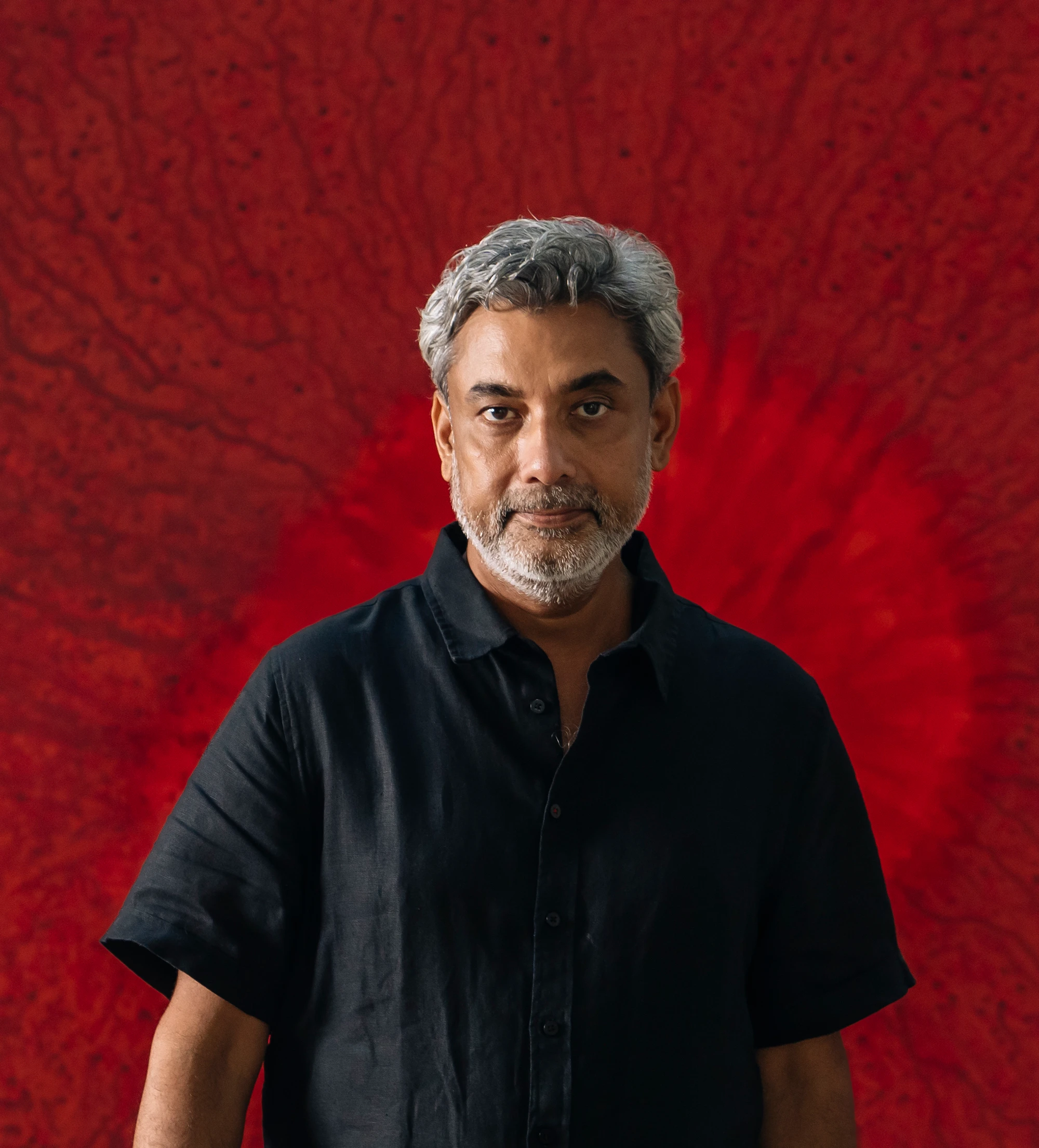 Indian contemporary artist Alwar Balasubramaniam standing before a deep red artwork background