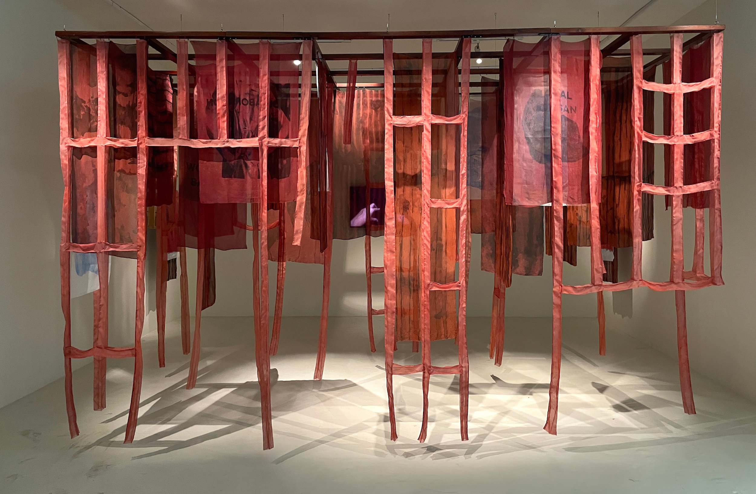 Dian Suci installation with red-dyed silk fabric panels hanging from teakwood frame, 2025